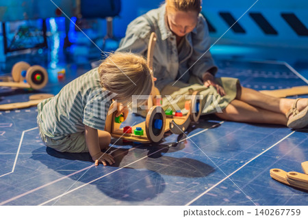 Mother sitting on floor with three year old toddler while building wooden toy machine at science center, hands-on learning and family education concept Mother sitting on floor with three year old toddler while building wooden toy machine at science center, hands-on learning and family education concept 140267759