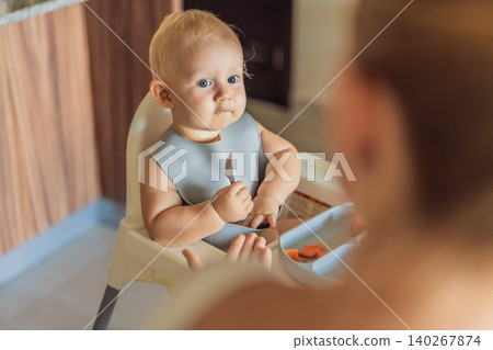 Infant practices self feeding with vegetables in a kitchen, showing baby led weaning, early nutrition and family care concept Infant practices self feeding with vegetables in a kitchen, showing baby led weaning, early nutrition and family care concept 140267874