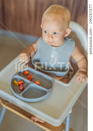Infant practices self feeding with vegetables in a kitchen, showing baby led weaning, early nutrition and family care concept Infant practices self feeding with vegetables in a kitchen, showing baby led weaning, early nutrition and family care concept 140267877