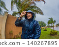 Adult man faces heavy rain and strong wind near palm trees and city buildings, showing storm weather, hurricane season and climate resilience concept 140267895