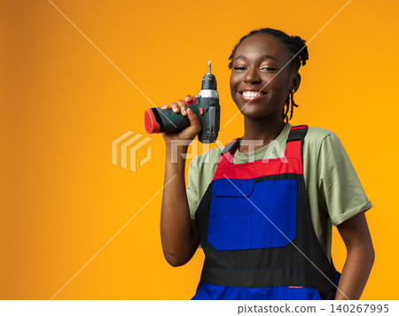 Young person holds a drill and smiles against a bright orange background in a workshop setting 140267995