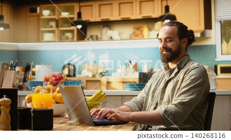 Man working remotely from his home setup in the kitchen, focused on solving daily administrative tasks using a laptop and wireless internet. Digital nomad lifestyle, flexible work routine. Man working remotely from his home setup in the kitchen, focused on solving daily administrative tasks using a laptop and wireless internet. Digital nomad lifestyle, flexible work routine. 140268116