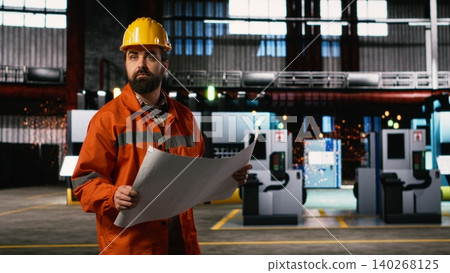 Engineer walking along facility reviewing manufacturing blueprints and technical plans, heavy industry warehouse. Production hall features machinery instructions, layout drawings. Engineer walking along facility reviewing manufacturing blueprints and technical plans, heavy industry warehouse. Production hall features machinery instructions, layout drawings. 140268125