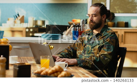 Soldier in military uniform spending quiet morning in the kitchen, sitting with laptop and coffee cup. Off duty relaxation, domestic life and everyday routine, calm moments away from active duty. Soldier in military uniform spending quiet morning in the kitchen, sitting with laptop and coffee cup. Off duty relaxation, domestic life and everyday routine, calm moments away from active duty. 140268133