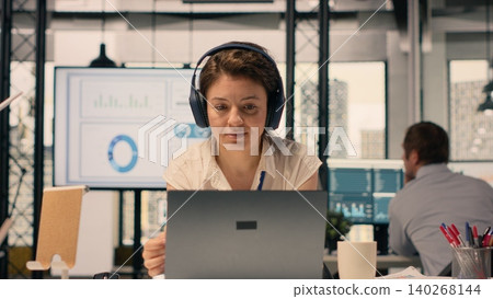 Businesswoman listening to music wearing wireless headphones, working in modern office. Relaxed multitasking moment with woman employee using laptop, combining stress relief with productivity. Businesswoman listening to music wearing wireless headphones, working in modern office. Relaxed multitasking moment with woman employee using laptop, combining stress relief with productivity. 140268144