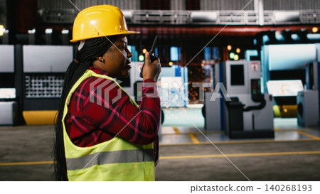 Black woman engineer speaking into portable walkie-talkie device, walking through warehouse and monitoring heavy equipment. Large-scale industrial facility highlights production line. 140268193