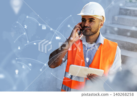 Logistics worker in safety gear uses phone and tablet at a construction site during daytime Logistics worker in safety gear uses phone and tablet at a construction site during daytime 140268258