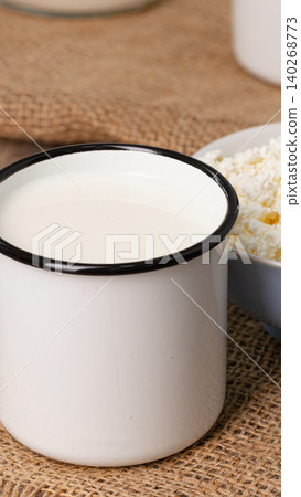 Fresh milk in a white cup beside a bowl of cheese on a burlap surface 140268773