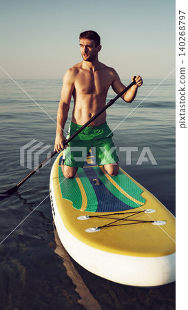 Man paddles on a stand-up paddleboard in calm water during sunset near the shore Man paddles on a stand-up paddleboard in calm water during sunset near the shore 140268797
