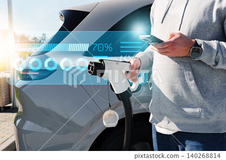 Man charges electric car at a station while checking phone in urban area during daylight Man charges electric car at a station while checking phone in urban area during daylight 140268814