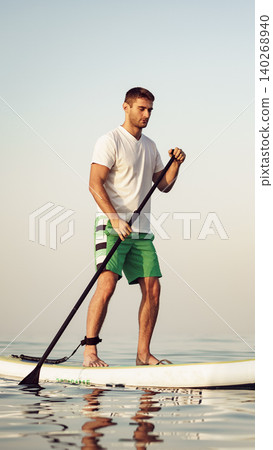 Man stands on paddleboard in calm water at sunrise near a beach Man stands on paddleboard in calm water at sunrise near a beach 140268940