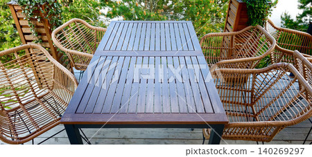 Outdoor patio dining set featuring a wooden table with slatted surface and four rattan chairs surrounded by greenery on a balcony Outdoor patio dining set featuring a wooden table with slatted surface and four rattan chairs surrounded by greenery on a balcony 140269297