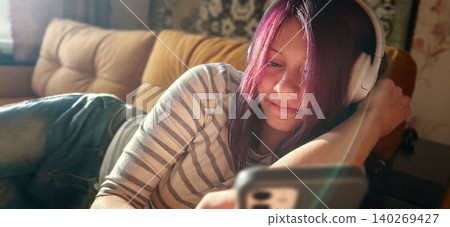 Teenage girl with purple hair listens to music on smartphone while lounging on a couch at home, wearing headphones in a cozy living room setting Teenage girl with purple hair listens to music on smartphone while lounging on a couch at home, wearing headphones in a cozy living room setting 140269427