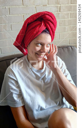 Teenage girl with a red towel on her head smiles while sitting on a couch in a cozy indoor setting with a light-colored brick wall behind her Teenage girl with a red towel on her head smiles while sitting on a couch in a cozy indoor setting with a light-colored brick wall behind her 140269434