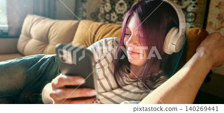 Young woman with purple hair wearing headphones relaxes on a couch while using a smartphone, with a cozy living room setting in the background Young woman with purple hair wearing headphones relaxes on a couch while using a smartphone, with a cozy living room setting in the background 140269441