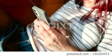 Teen girl with red hair using smartphone while sitting on a couch, wearing a striped shirt and denim shorts, sunlight illuminating the scene Teen girl with red hair using smartphone while sitting on a couch, wearing a striped shirt and denim shorts, sunlight illuminating the scene 140269453