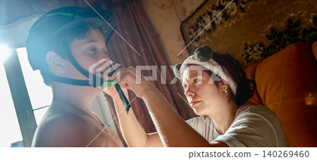 Sister helps younger brother fasten bicycle helmet indoors, with soft lighting and patterned wall decor creating a cozy atmosphere in the background Sister helps younger brother fasten bicycle helmet indoors, with soft lighting and patterned wall decor creating a cozy atmosphere in the background 140269460