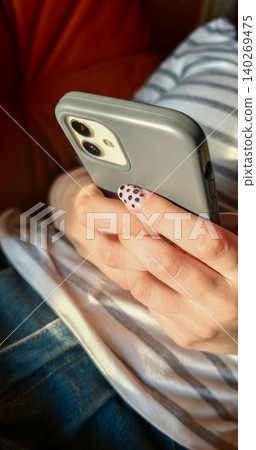 Close-up of a female hand holding a smartphone with a gray case, featuring polka dot nail art, resting on a striped fabric couch in a cozy indoor setting Close-up of a female hand holding a smartphone with a gray case, featuring polka dot nail art, resting on a striped fabric couch in a cozy indoor setting 140269475