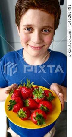 Young boy with brown hair wearing blue shirt holds yellow plate filled with fresh strawberries in a bright kitchen setting with modern decor Young boy with brown hair wearing blue shirt holds yellow plate filled with fresh strawberries in a bright kitchen setting with modern decor 140269481