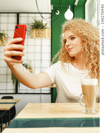 Woman takes selfie in cafe while enjoying coffee in a bright green space Woman takes selfie in cafe while enjoying coffee in a bright green space 140270440