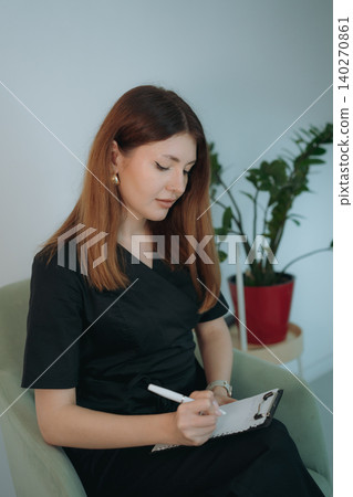 young woman dressed in black medical suit holding a clipboard seated on armchair in modern clinic young woman dressed in black medical suit holding a clipboard seated on armchair in modern clinic 140270861
