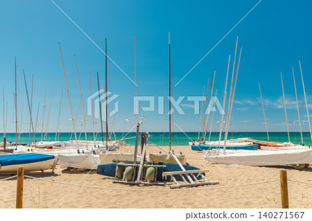 Sailing boats and catamarans stored on a sandy Mediterranean beach in Spain. Turquoise sea, clear blue sky and yacht club facilities Sailing boats and catamarans stored on a sandy Mediterranean beach in Spain. Turquoise sea, clear blue sky and yacht club facilities 140271567