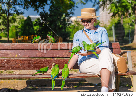 Woman feeding colorful monk parakeets while sitting on park bench in Barcelona, friendly green tropical birds gathering around tourist outdoors Woman feeding colorful monk parakeets while sitting on park bench in Barcelona, friendly green tropical birds gathering around tourist outdoors 140271680