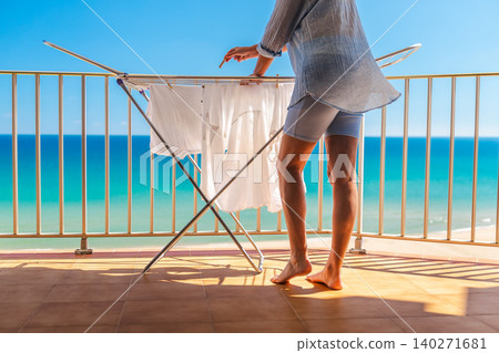 Woman hanging white laundry on drying rack on sunny balcony with turquoise sea view during summer vacation by the beach Woman hanging white laundry on drying rack on sunny balcony with turquoise sea view during summer vacation by the beach 140271681