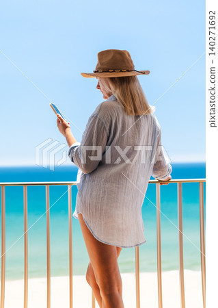 Woman in sheer beach cover up and cowboy hat using smartphone on seaside balcony, tropical summer vacation and remote lifestyle concept Woman in sheer beach cover up and cowboy hat using smartphone on seaside balcony, tropical summer vacation and remote lifestyle concept 140271692