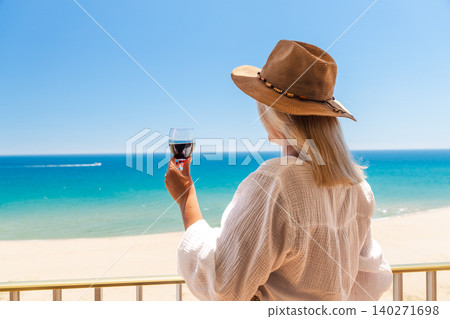 Woman in straw hat holding glass of red wine on balcony with turquoise sea and sandy beach during luxury summer vacation 140271698