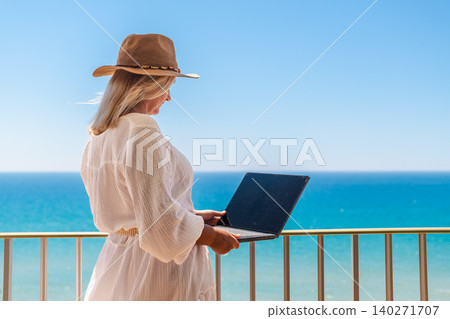 Woman working remotely on laptop from seaside balcony with ocean view during relaxing summer vacation at beach resort Woman working remotely on laptop from seaside balcony with ocean view during relaxing summer vacation at beach resort 140271707