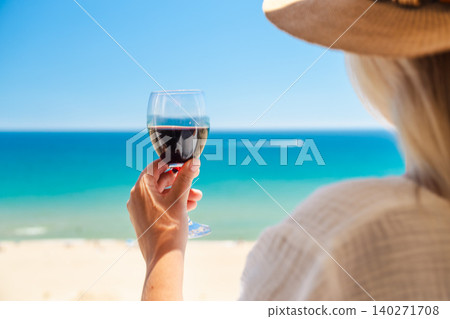 Close up of woman holding glass of red wine with turquoise sea and sandy beach in background during luxury summer vacation Close up of woman holding glass of red wine with turquoise sea and sandy beach in background during luxury summer vacation 140271708