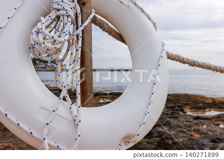 White lifebuoy rescue ring with rope on a rocky Mediterranean seafront in Paphos, Cyprus, symbolizing coastal water safety and emergency preparedness. White lifebuoy rescue ring with rope on a rocky Mediterranean seafront in Paphos, Cyprus, symbolizing coastal water safety and emergency preparedness. 140271899