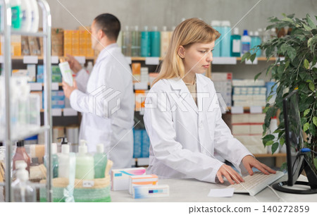 Young woman working with computer and male pharmacist arranging shelves behind Young woman working with computer and male pharmacist arranging shelves behind 140272859