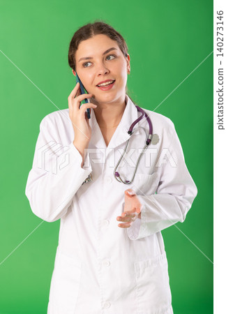 Young female doctor in white coat with stethoscope talking on mobile phone on green background 140273146