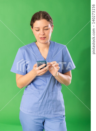 Portrait of polite female doctor wearing blue medical clothing scrolling smartphone standing against green screen Portrait of polite female doctor wearing blue medical clothing scrolling smartphone standing against green screen 140273156
