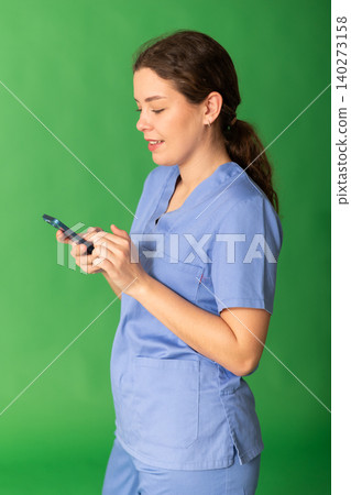 Portrait of polite female doctor wearing blue medical clothing scrolling smartphone standing against green screen Portrait of polite female doctor wearing blue medical clothing scrolling smartphone standing against green screen 140273158