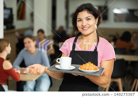 Positive female waitress carries coffee and croissants on tray Positive female waitress carries coffee and croissants on tray 140273307