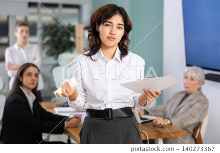 Sad Asian girl with documents stands at distance from office staff Sad Asian girl with documents stands at distance from office staff 140273367