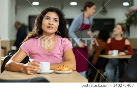 Young Latina girl enjoying coffee and croissant in cafeteria Young Latina girl enjoying coffee and croissant in cafeteria 140273403