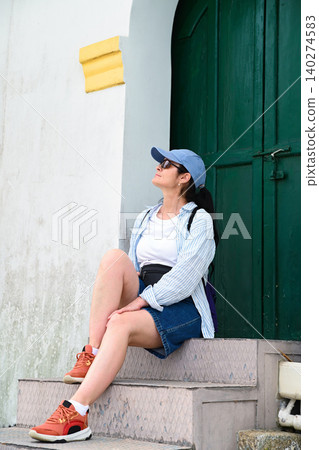 Close up of mature 50 years old Russian woman tourist in cap sitting on stairs near ancient green door of Guia Lighthouse in Macau destination Close up of mature 50 years old Russian woman tourist in cap sitting on stairs near ancient green door of Guia Lighthouse in Macau destination 140274583