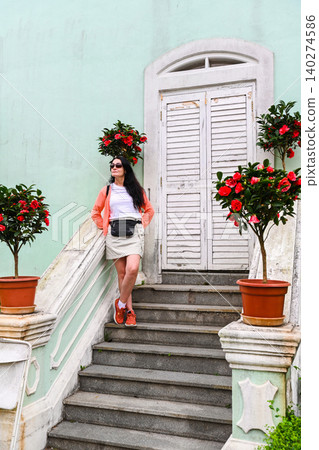 Mature 50 years old Russian woman tourist in cap standing on stairs near mint green house built in 1921 with camellia flowers in Taipa Houses Macau Mature 50 years old Russian woman tourist in cap standing on stairs near mint green house built in 1921 with camellia flowers in Taipa Houses Macau 140274586