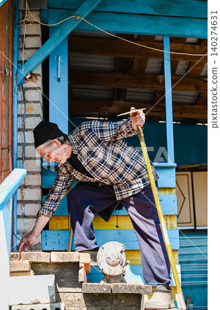 Russian 77 years old elderly man holding hoe tool intends to sharpen it on vintage electric grinding machine near house porch in village Russian 77 years old elderly man holding hoe tool intends to sharpen it on vintage electric grinding machine near house porch in village 140274631