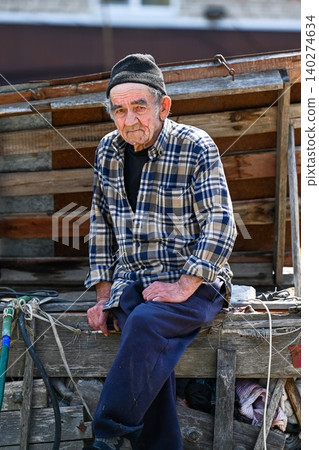 Poor russian 77 years old elderly man sits outdoors looking into camera showing concept of poverty in rural village Poor russian 77 years old elderly man sits outdoors looking into camera showing concept of poverty in rural village 140274634