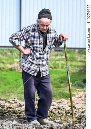 Tired russian 77 years old elderly man leans on shovel holding his aching lower back while digging soil during potato planting season in rural village Tired russian 77 years old elderly man leans on shovel holding his aching lower back while digging soil during potato planting season in rural village 140274635