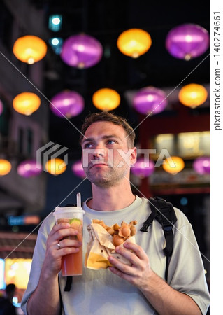 Handsome 30 years old Russian man tourist chewing traditional bubble waffle and holding iced lemon tea at Temple Street Night Market in Hong Kong Handsome 30 years old Russian man tourist chewing traditional bubble waffle and holding iced lemon tea at Temple Street Night Market in Hong Kong 140274661