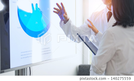 A group of men and women in white coats holding a meeting while looking at a screen. A group of men and women in white coats holding a meeting while looking at a screen. 140275559
