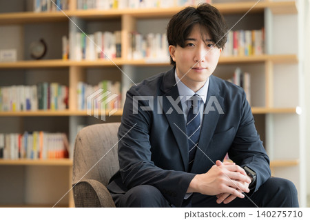 Portrait of a handsome young executive, CEO, or businessman sitting in front of a bookshelf, looking directly at the camera. Portrait of a handsome young executive, CEO, or businessman sitting in front of a bookshelf, looking directly at the camera. 140275710