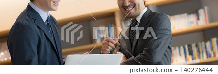 A business executive and his subordinate are seen smiling while having a meeting in front of a bookshelf resembling a CEO's office, looking at their computers. (Faceless) A business executive and his subordinate are seen smiling while having a meeting in front of a bookshelf resembling a CEO's office, looking at their computers. (Faceless) 140276444