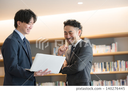 A smiling businessman and his boss are having a meeting in front of a bookshelf that looks like a CEO's office, both looking at their computers. 140276445